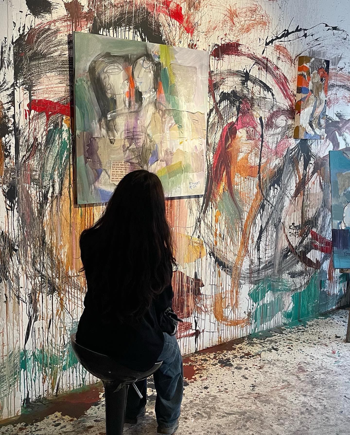 Person seated before layered abstract paintings in a room covered with energetic red, black, teal, and ochre marks.