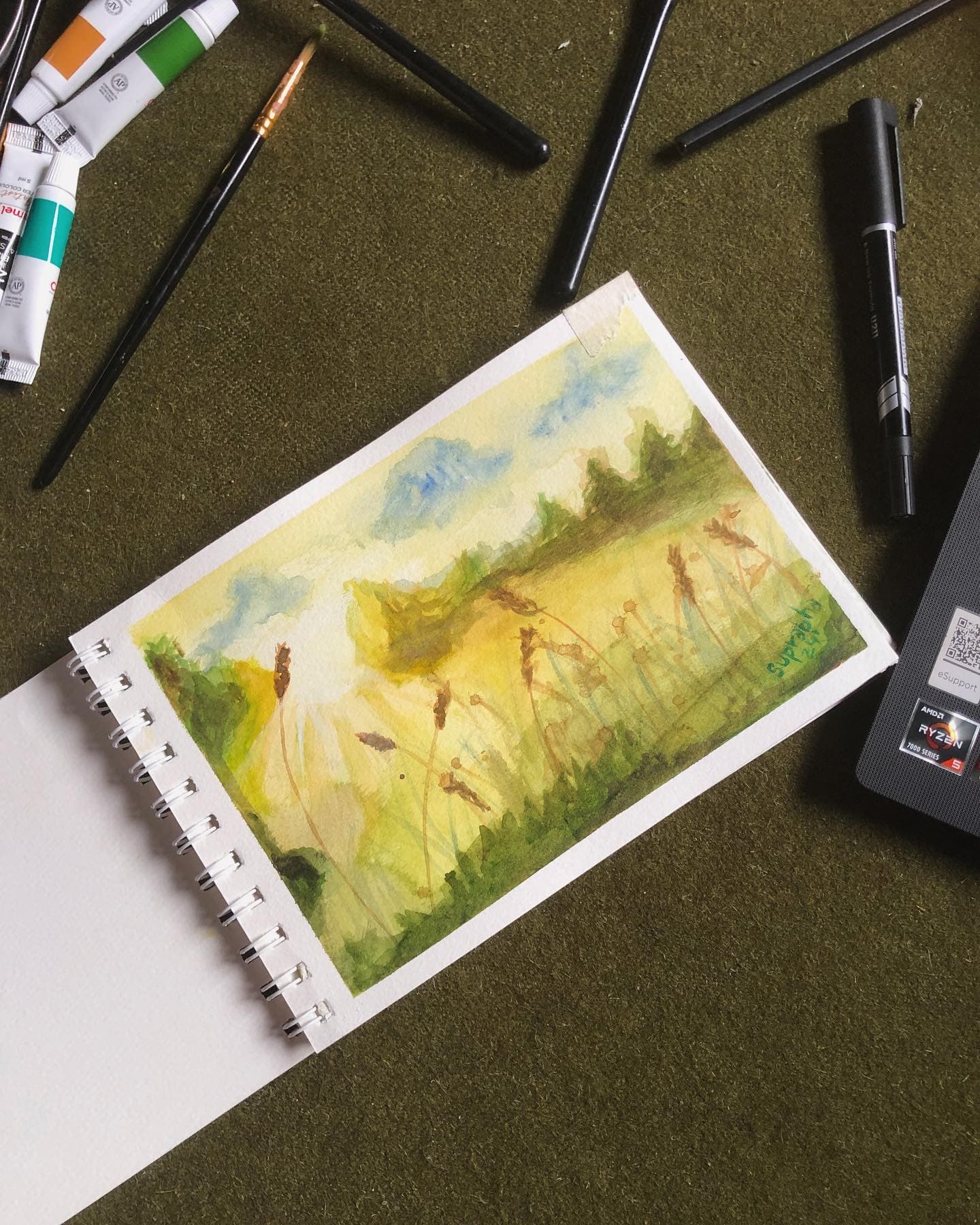 Small watercolor of a sunlit yellow-green field and distant trees, open on a desk among art tools.
