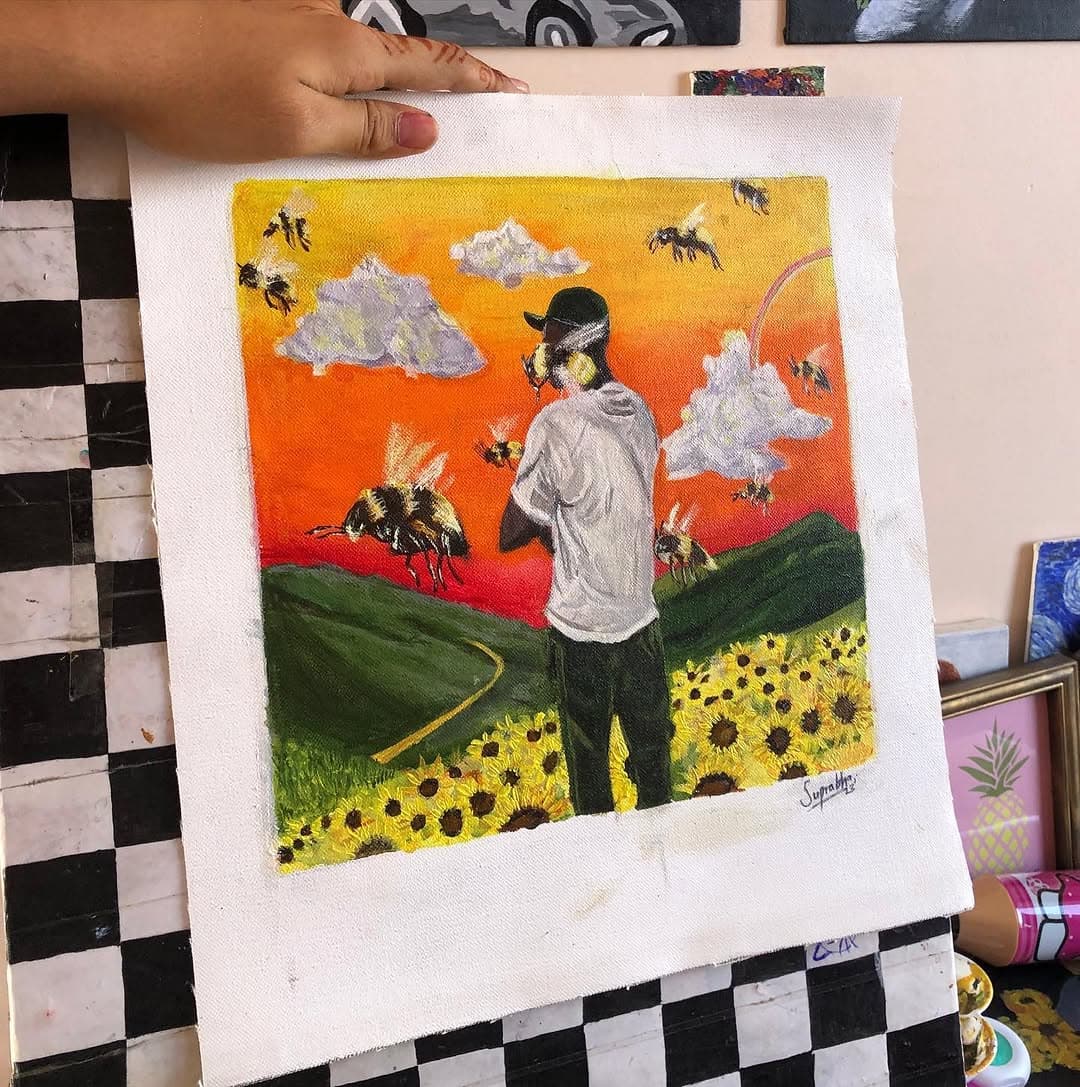 A hand holds a canvas showing a lone figure among yellow sunflowers and giant bees beneath a blazing orange sky.