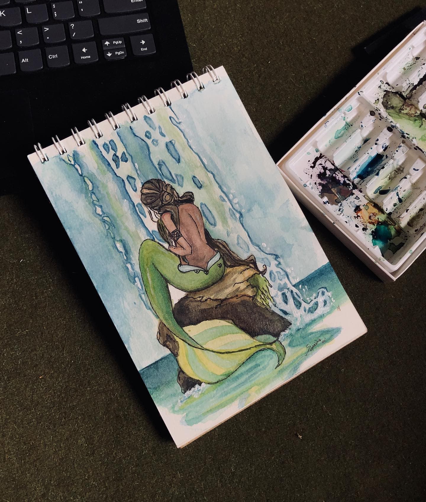 Watercolor sketchbook illustration of a curled seated mermaid surrounded by loose blue water marks.