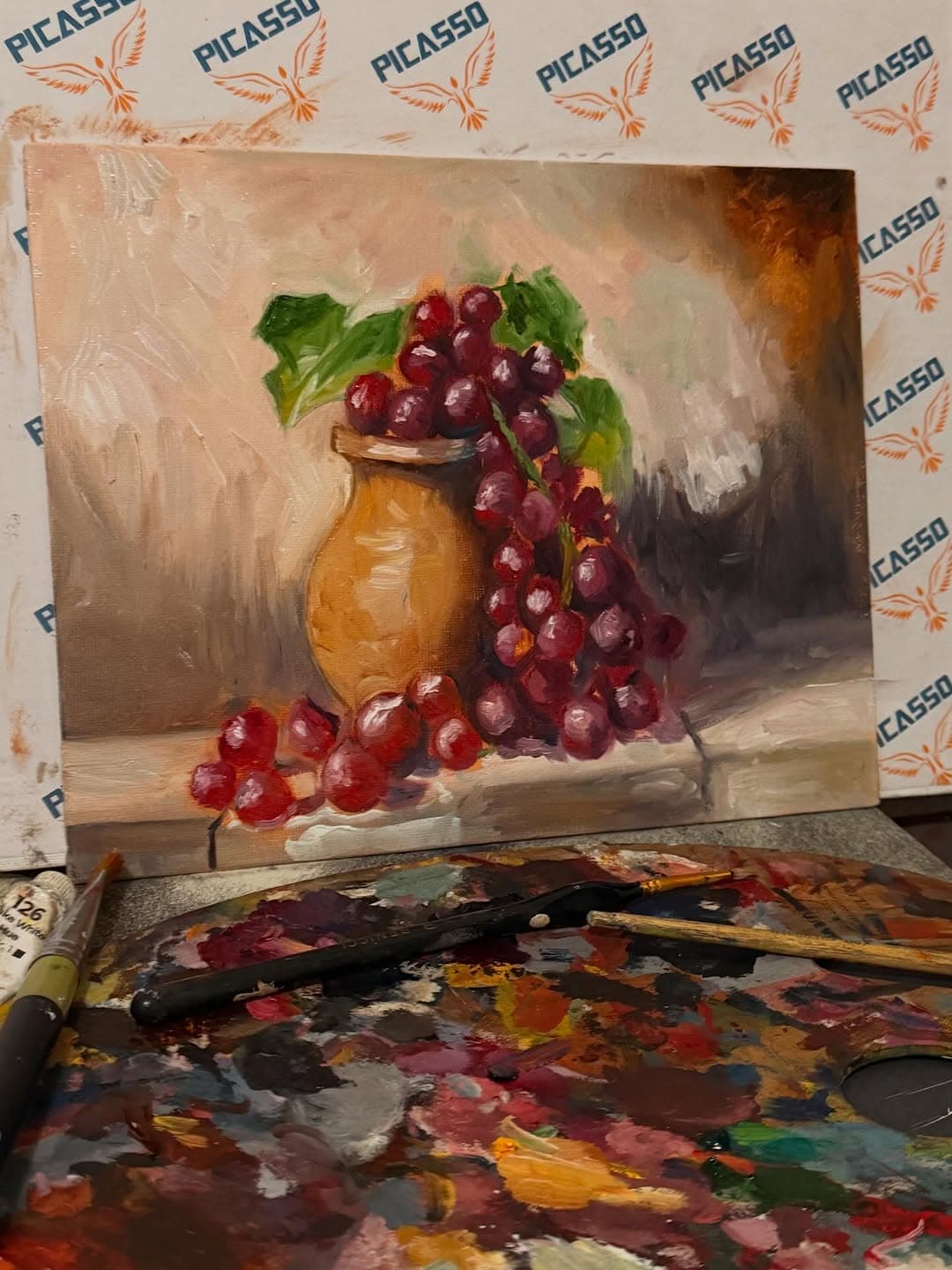 Still-life painting of burgundy grapes spilling from an ochre vase above a heavily used palette and brushes.