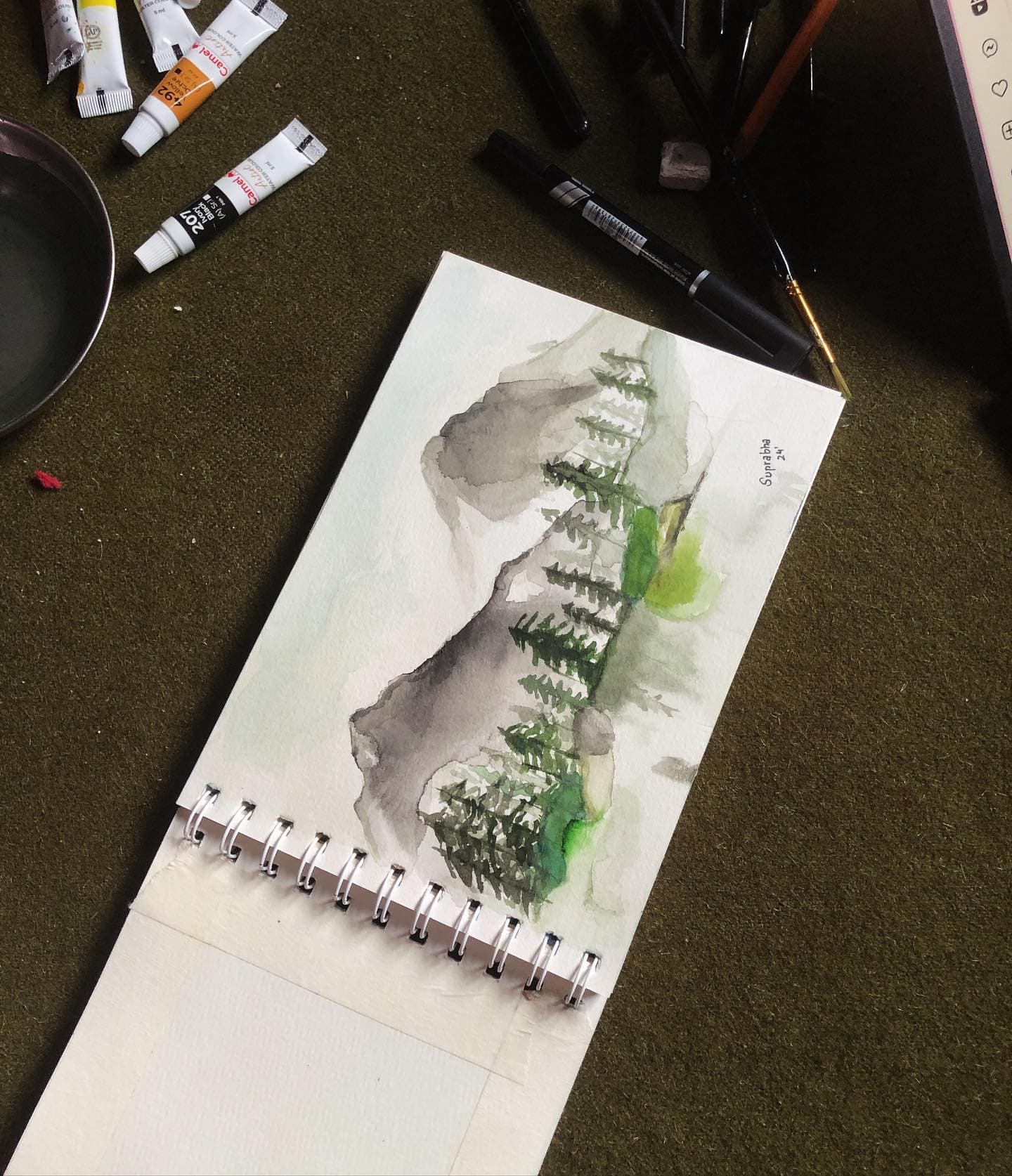 Vertical watercolor landscape of misty dark trees with a small bright green passage, surrounded by pens and paint tubes.