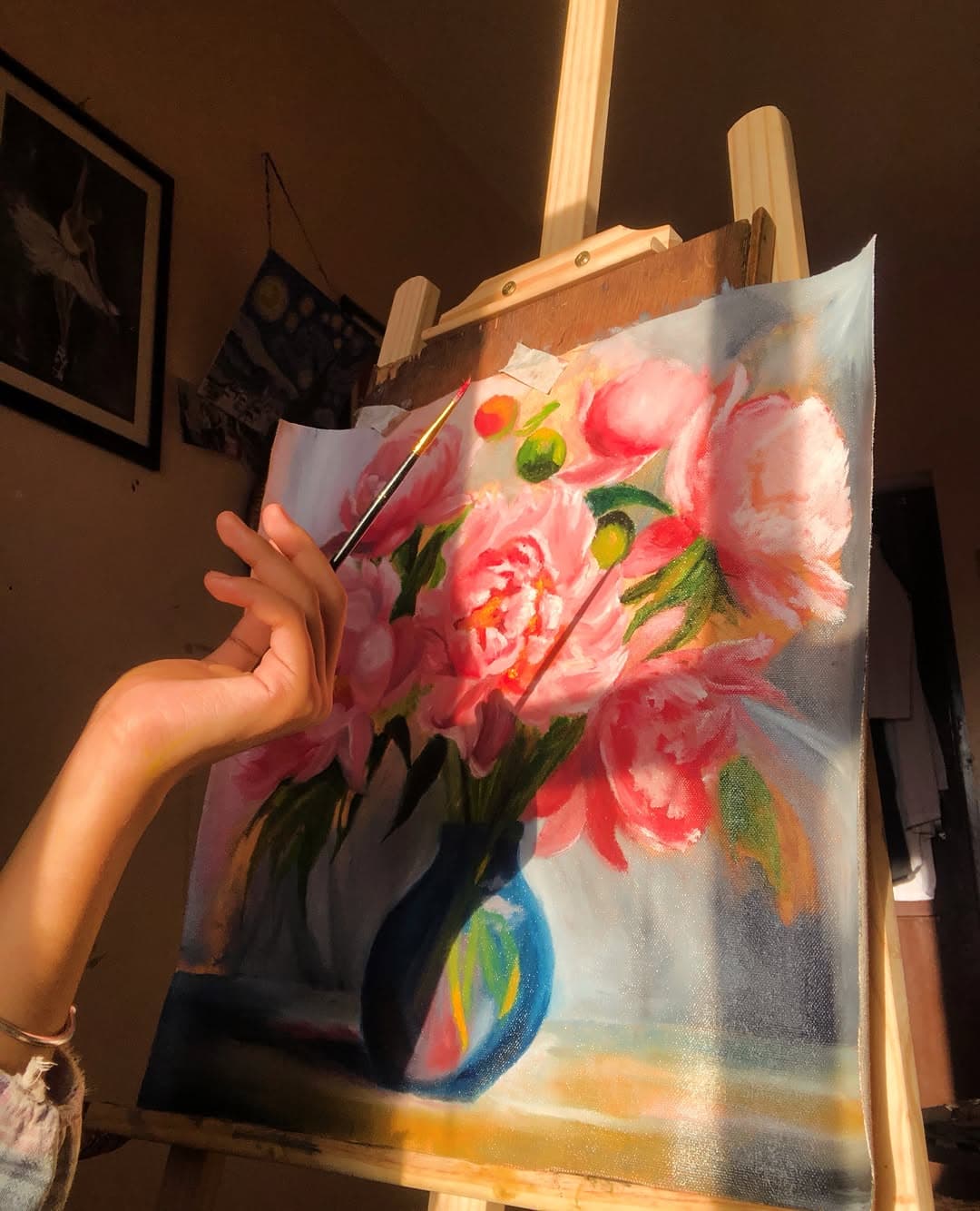 A hand paints pink flowers on a canvas at an easel as golden sunlight falls across the bouquet and blue vase.