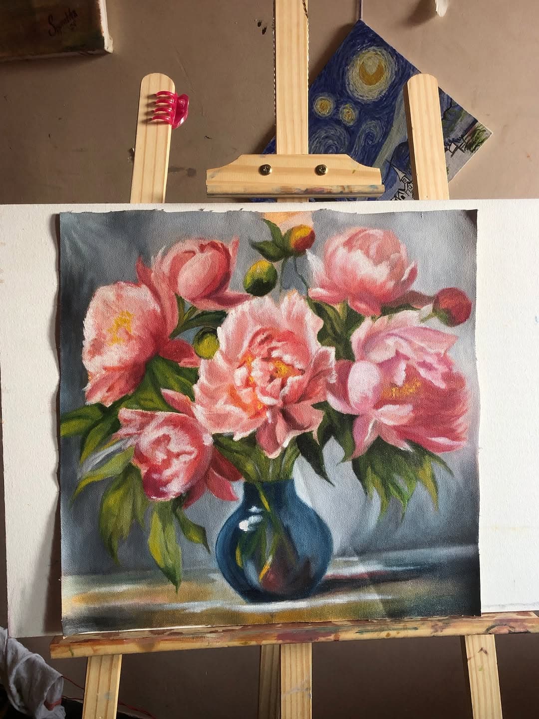 Painting of loose pink peonies in a dark blue vase displayed on a small easel.