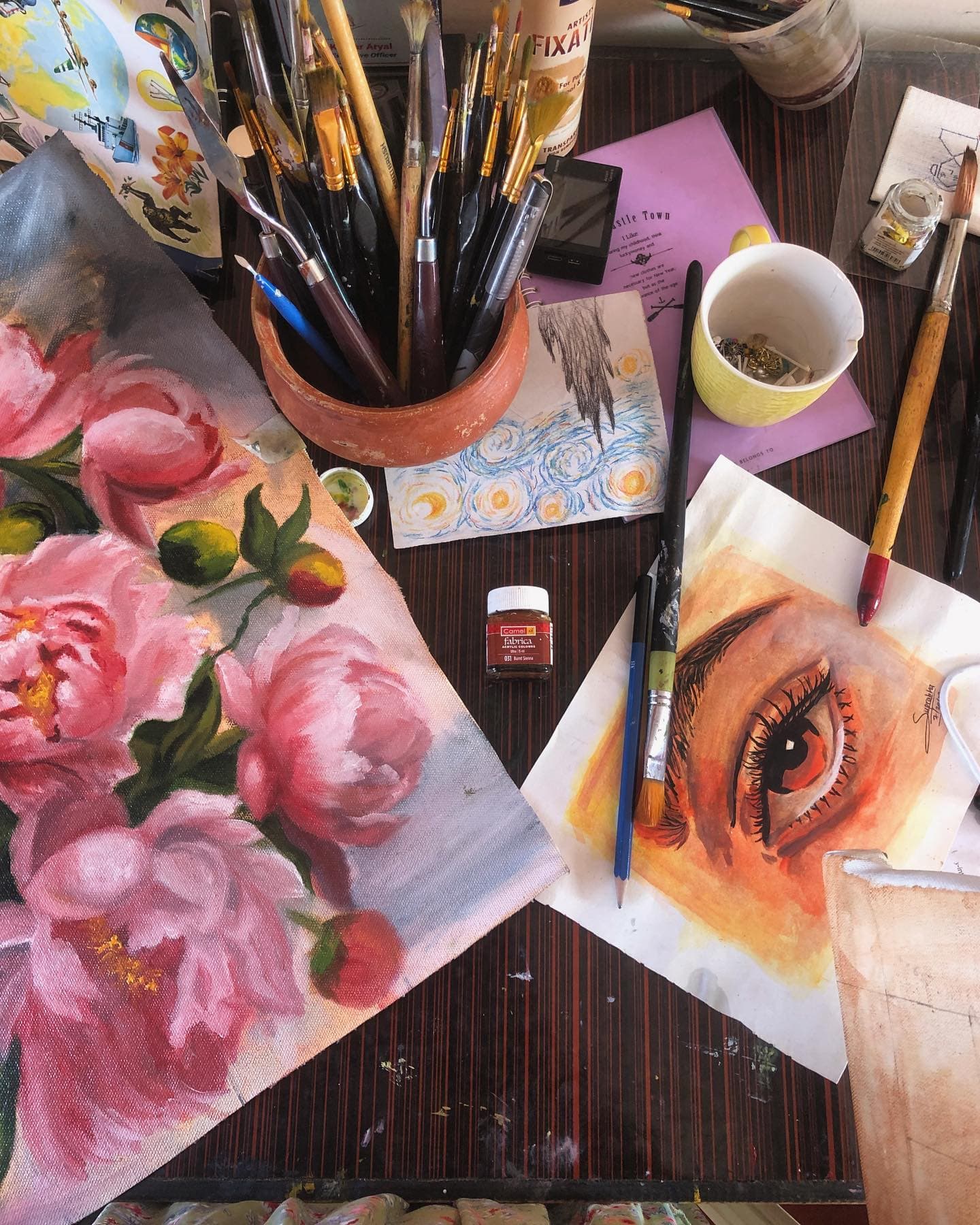Crowded artist's table with brushes, painted pink roses, an eye study, pigments, and a cup of tea.
