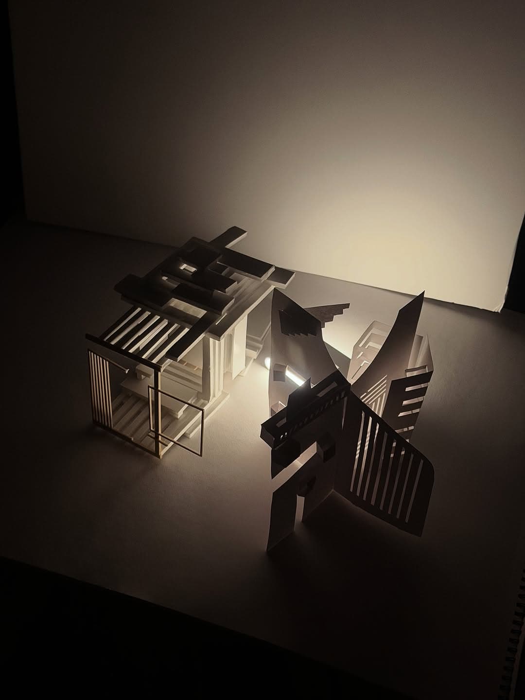 A fragmented black-and-white paper model glows from small interior lights, throwing angular shadows across a dark tabletop.
